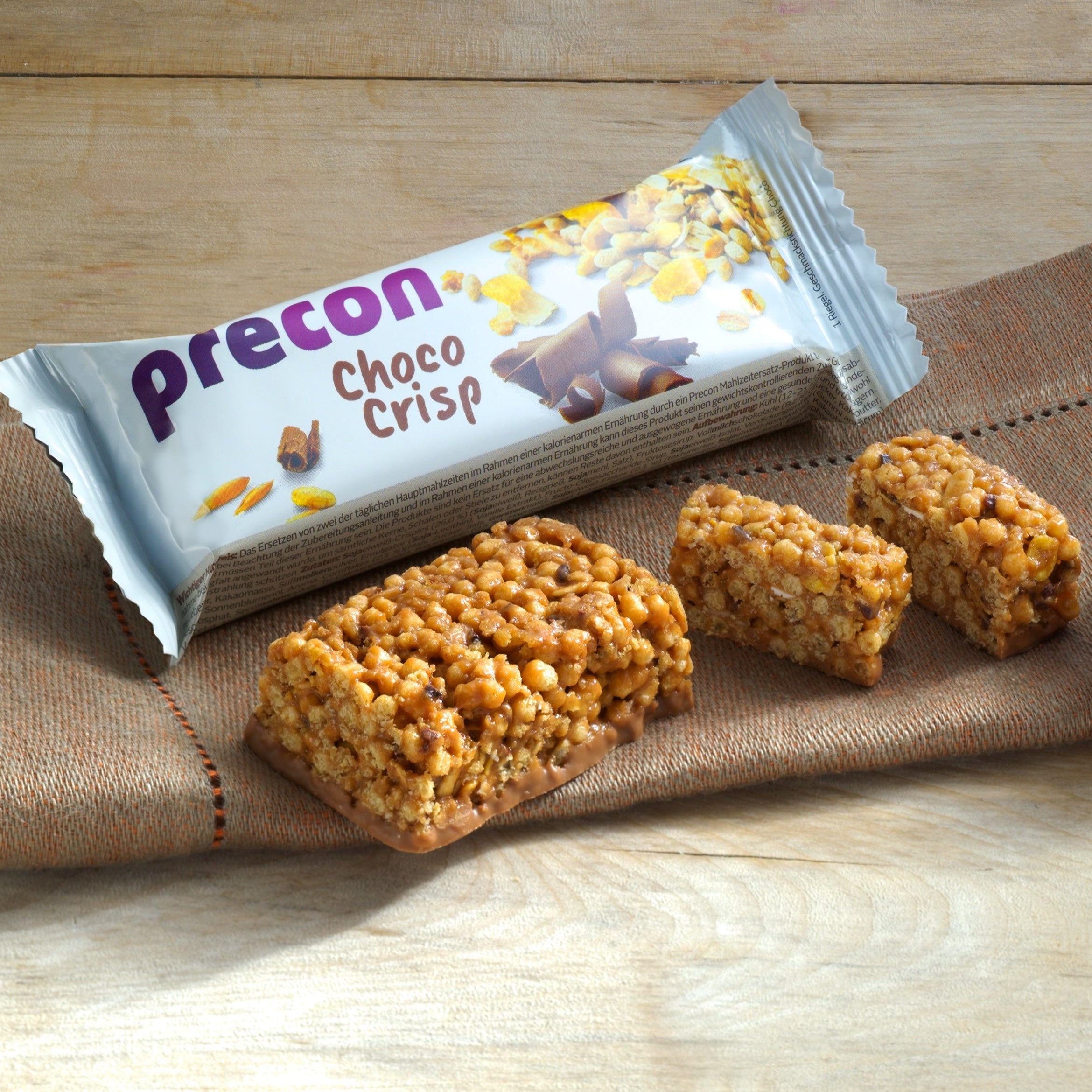 Ein Precon Riegel-Probier-Paket Choco Crisp wird auf einer hölzernen Oberfläche gezeigt, mit drei rechteckigen Mahlzeitenersatz-Stücken davor, die ihre knusprige Textur und ihren Schokoladenüberzug hervorheben.