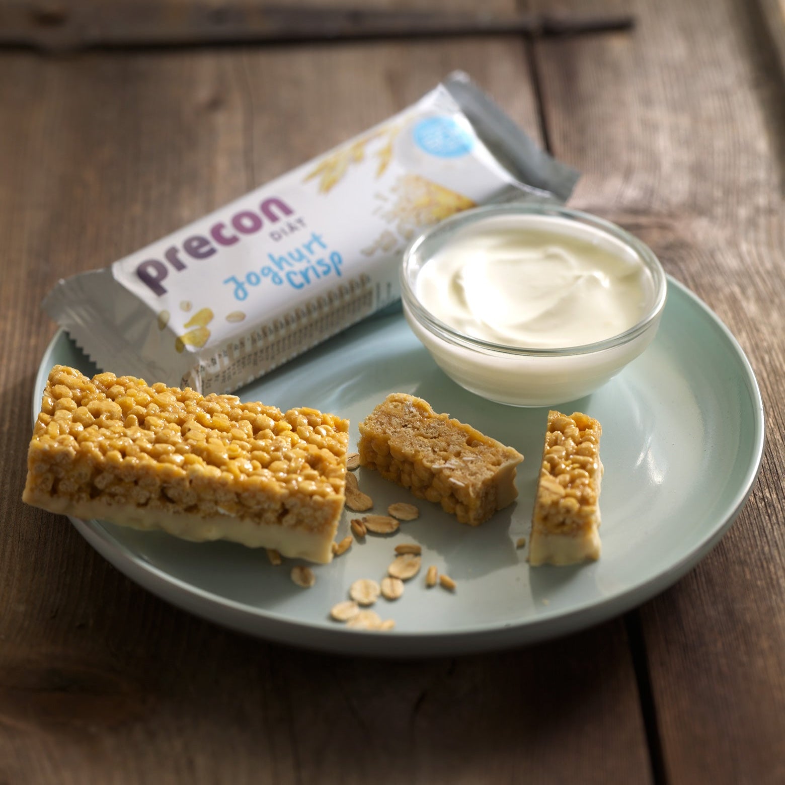 Ein Teller mit einem Precon JoghurtCrisp Riegel, zwei kleineren Stücken, einem Schälchen Joghurt und Haferflocken steht auf einem Holztisch mit der Snackbar-Verpackung im Hintergrund - ideal als Mahlzeitenersatz.