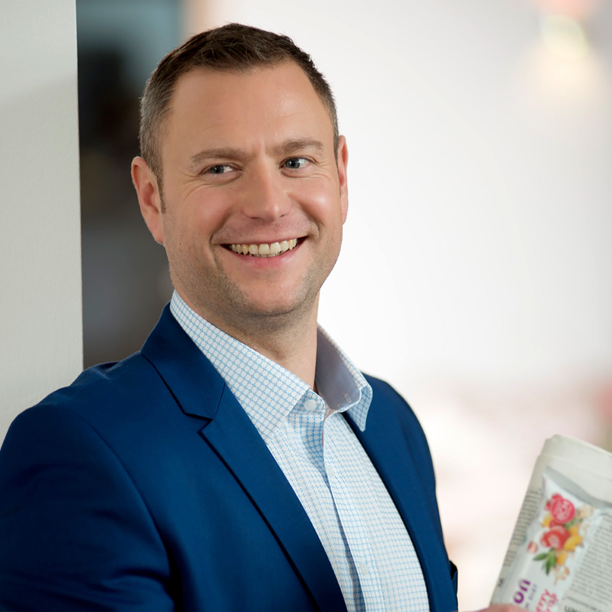 Ein lächelnder Mann in einer blauen Anzugsjacke und einem karierten Hemd steht in einem Haus und hält eine geblümte Tasse in der Hand, die er vielleicht mit seinem Lieblings-Precon CranberryCrisp Riegel als Teil seiner Mahlzeitersatzroutine genießt.