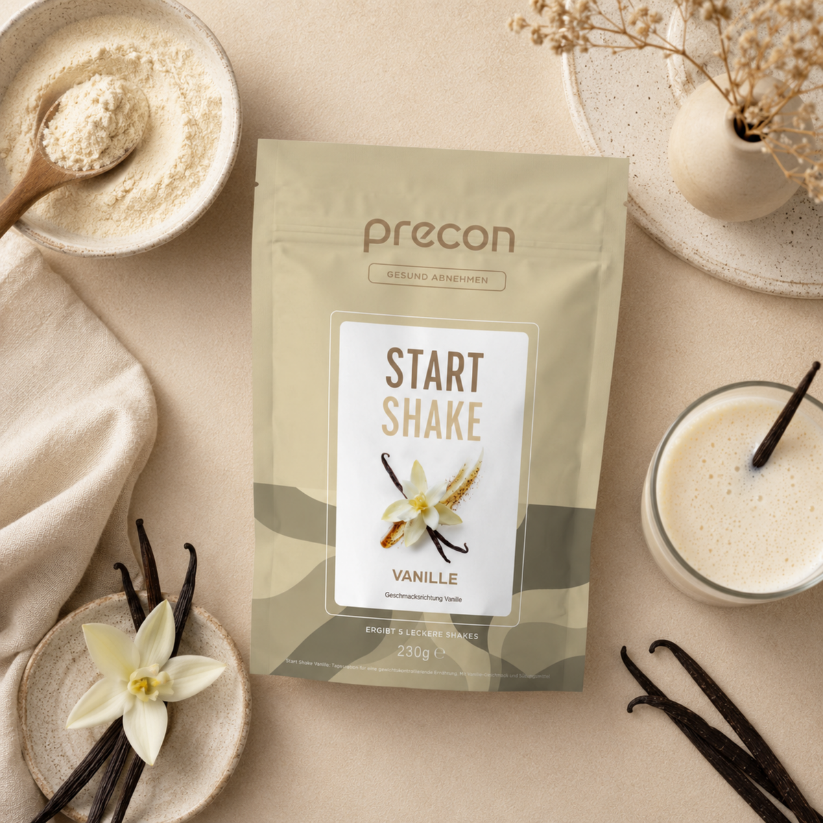 Ein „Start Shake Vanille“-Beutel der Anona GmbH liegt in einer Flat-Lay-Anordnung auf einem hellbraunen Tisch, daneben stehen ein Glas Vanille-Shake, eine Portion Pulver, Vanilleschoten, eine Vanilleblüte und getrocknete Blumen.