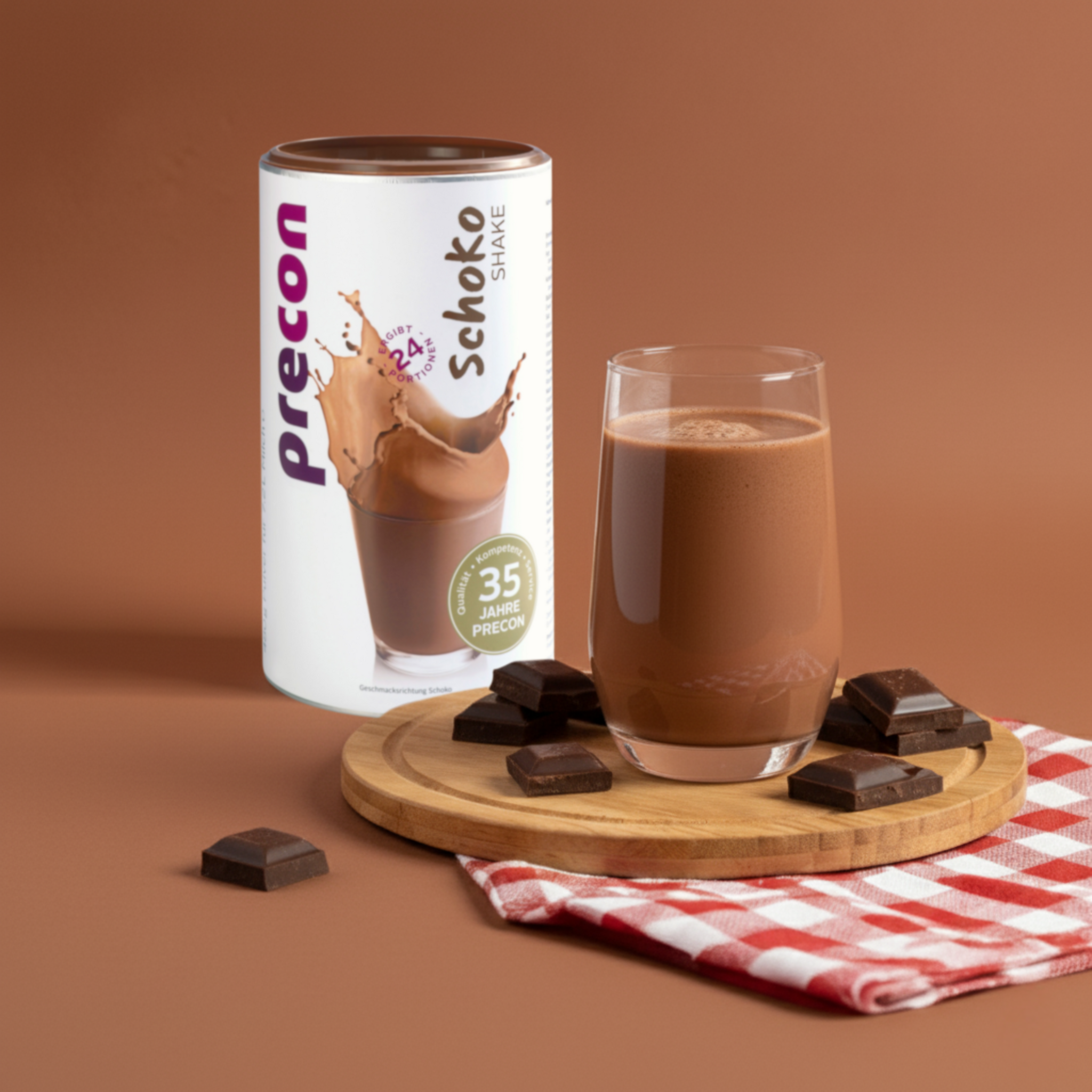 Ein Glas Precon Schoko Shake auf einem Holzbrett mit Schokoladenstückchen, eine rot-weiß karierte Serviette und eine Precon Schoko Shake Dose - ein köstlicher Mahlzeitenersatz zum Abnehmen - vor einem satten braunen Hintergrund.