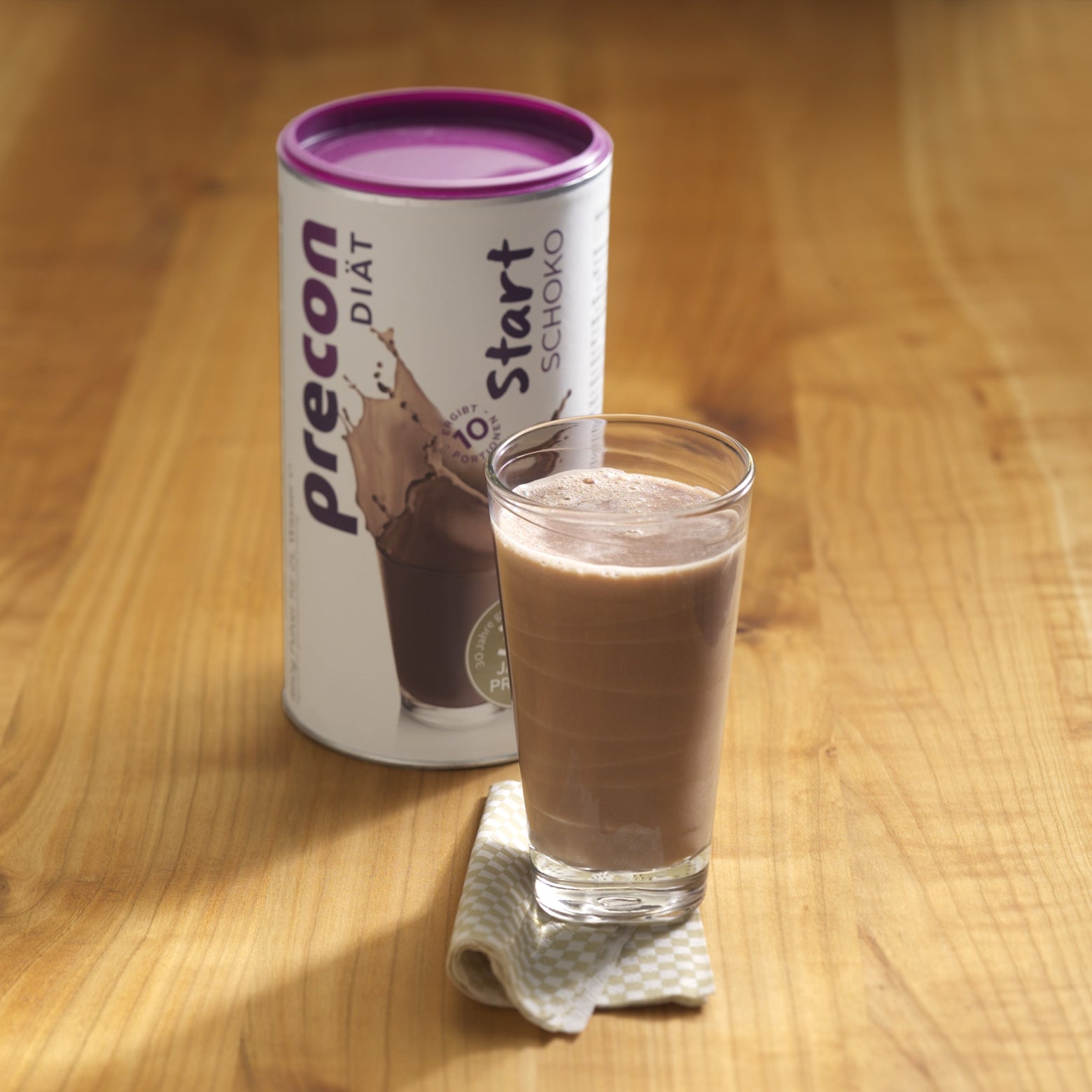 Ein Glas Schoko-Diät-Shake steht auf einer Serviette neben einem Precon Start Shake Schoko-Behälter - eine ideale Wahl für den Beginn einer Diät - auf einer Holzfläche.