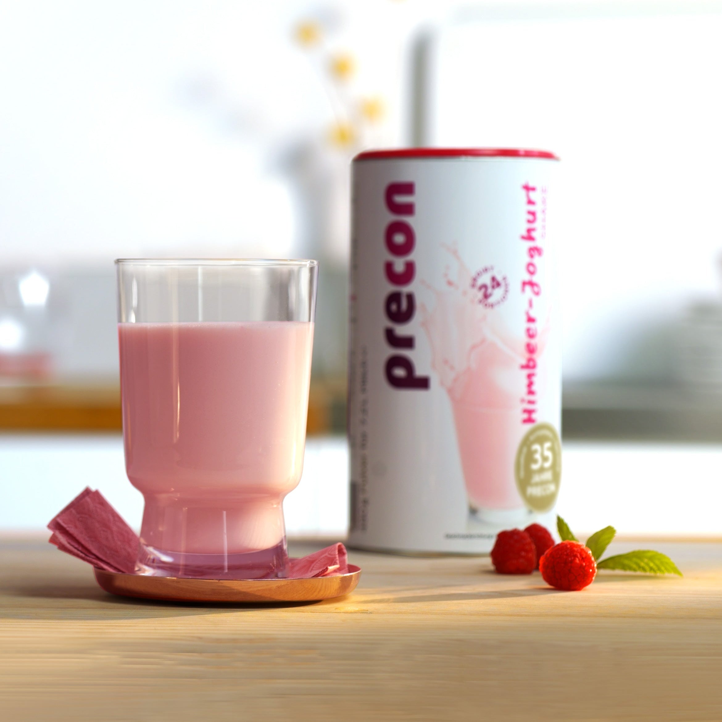 Auf einem Holztisch steht ein Glas mit rosa Himbeer-Joghurt-Getränk, daneben liegen eine Serviette und zwei Himbeeren. Dahinter steht ein Kanister Precon 1-Monats-Startpaket EU (ohne Handbuch), ein Top-Diätprodukt für diejenigen, die die Precon-Diät zur Unterstützung der Gewichtsabnahme machen.
