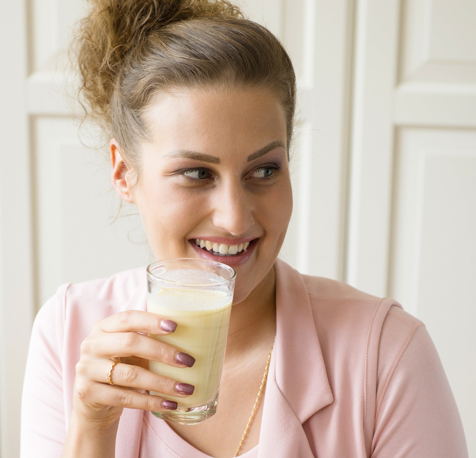 Eine Frau mit lockigem Haar in einem hellrosa Oberteil lächelt und hält ein Glas Precon Shake Mix - Einzelportionen in der rechten Hand. Sie blickt in einem hell erleuchteten Raum zur Seite und genießt ihren cremigen Mahlzeitenersatz.