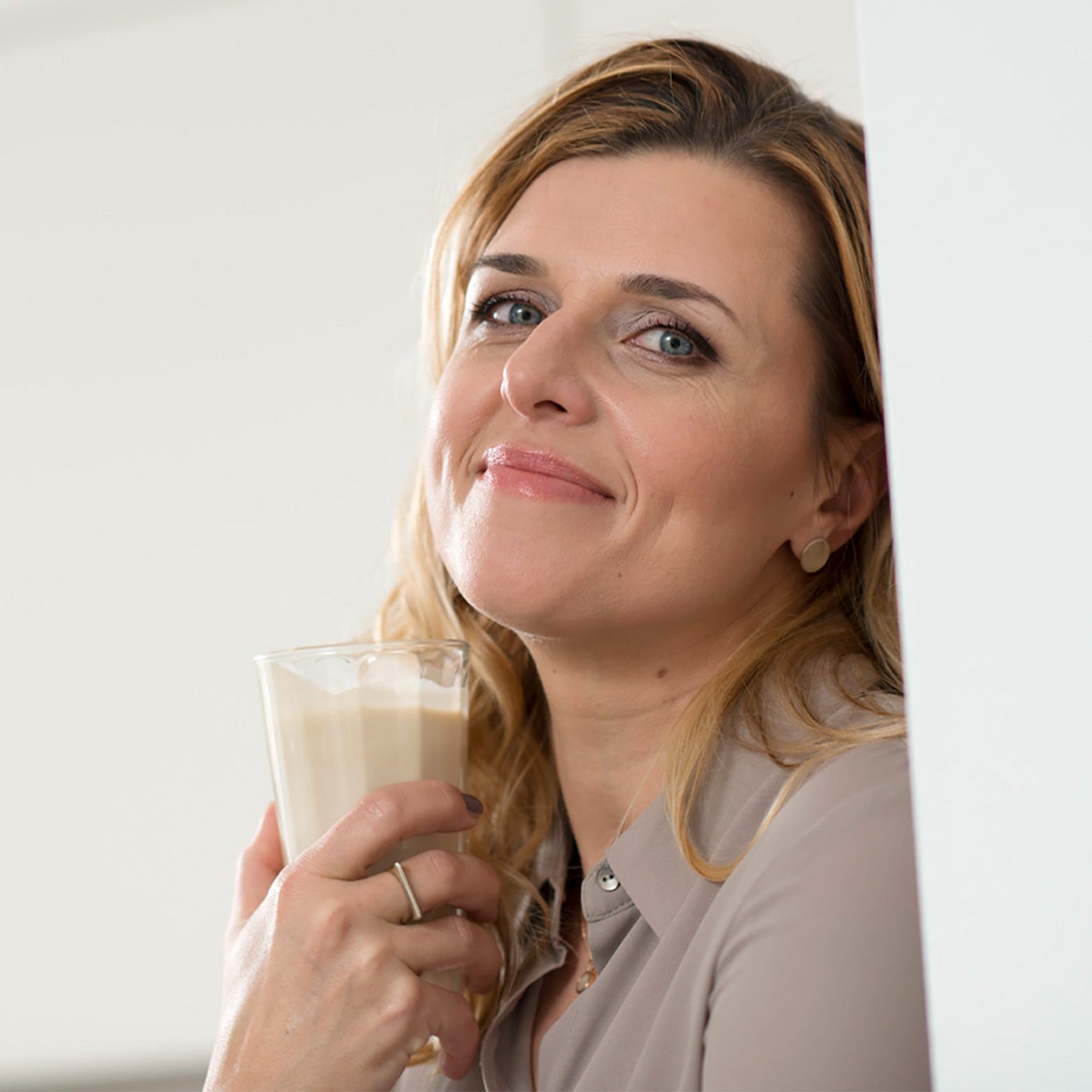 Eine Frau mit langem hellbraunem Haar lächelt sanft, während sie ein Glas mit cremigem Precon Abnehmshake aus ihrem Mix & Match Bundle dicht an ihr Gesicht hält. Sie steht in einem Innenraum und lehnt an einer weißen Wand.