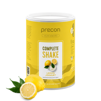Neben einer Zitrone und grünen Blättern steht ein gelber Behälter mit „Complete Shake“ der Anona GmbH in der Geschmacksrichtung Zitrone-Joghurt. Auf dem Etikett sind eine in Scheiben geschnittene Zitrone sowie die wichtigsten Nährwertangaben des Produkts abgebildet.