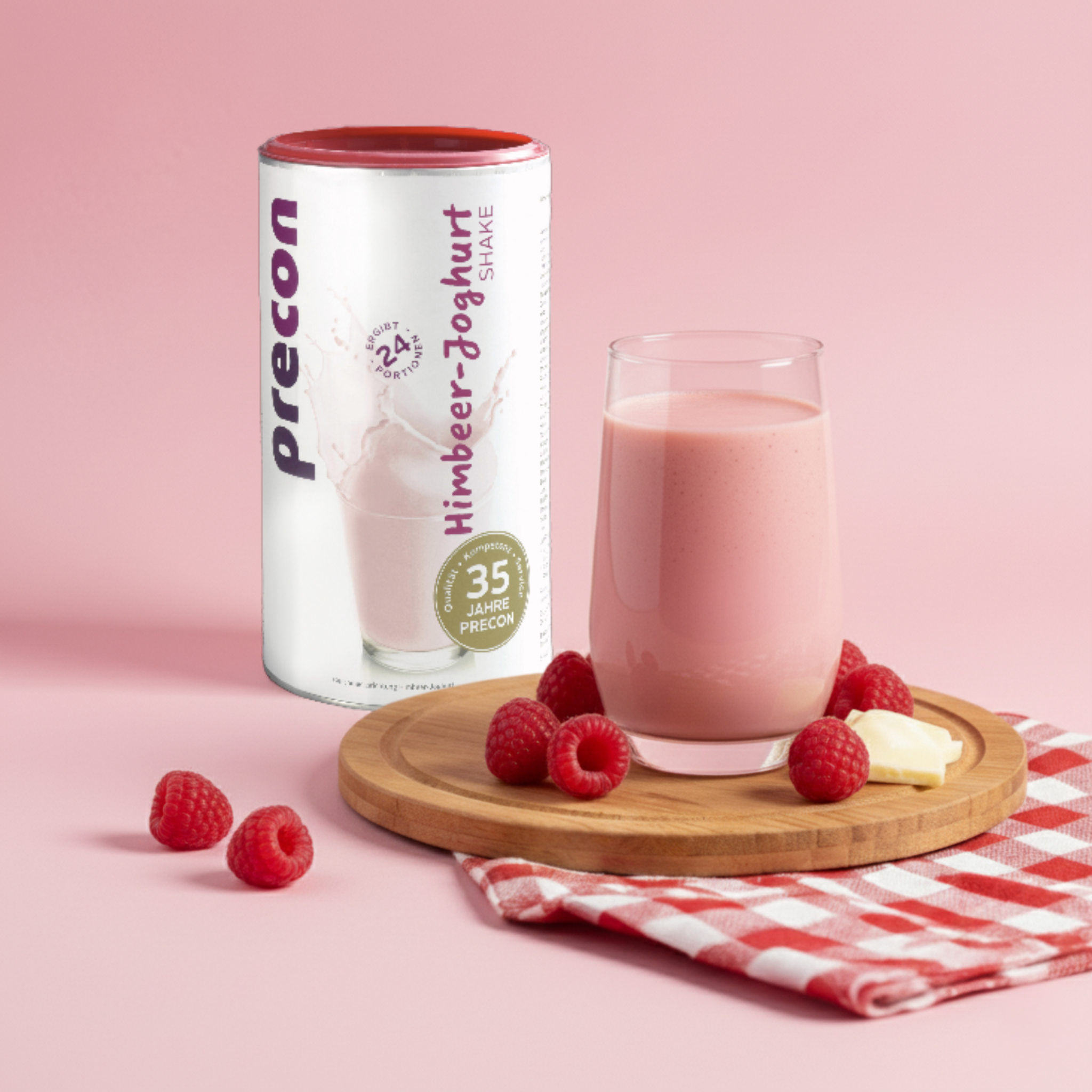 Ein Glas Precon Himbeer-Joghurt-Shake steht auf einem Holzbrett mit Himbeeren und weißer Schokolade, daneben der Behälter, alles auf einem rot-karierten Tuch vor einem rosa Hintergrund.