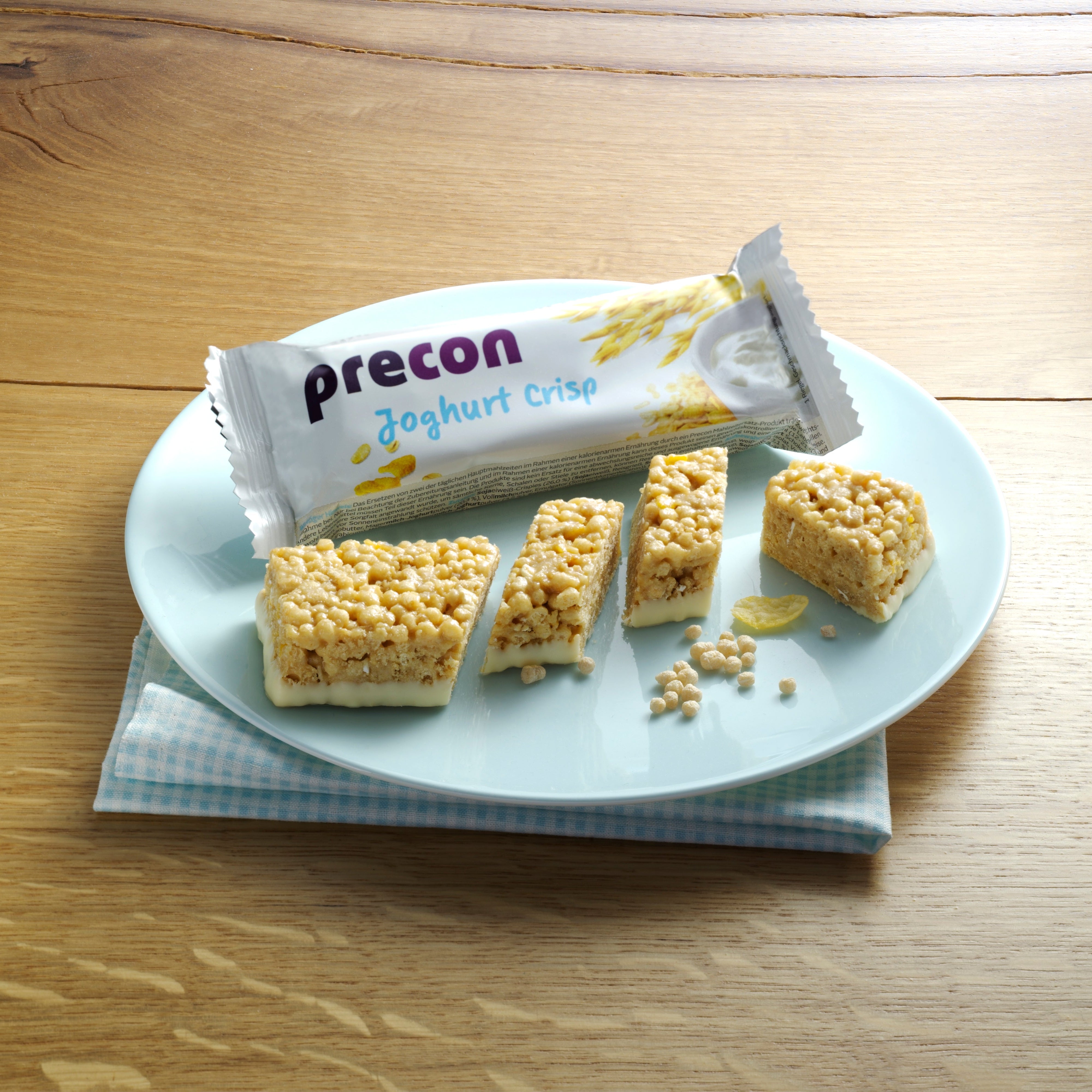 Ein hellblauer Teller mit einem Precon JoghurtCrisp Riegel, von dem einige Stücke abgeschnitten sind, liegt auf einer gefalteten, karierten Serviette. Die ungeöffnete Verpackung des Precon-Riegels liegt hinter den Stücken, die alle auf einer Holzfläche angeordnet sind.
